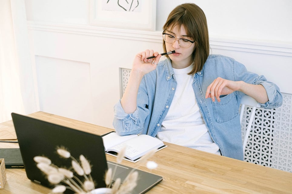 Thoughtful-woman-biting-on-her-pen-while-looking-at-notebook-and-laptop-pros-and-cons-list