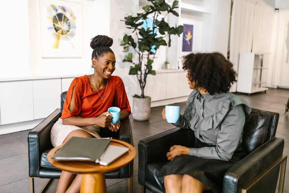 Woman-with-her-coach-talking-and-drinking-coffee-at-lounge-interview-coaching